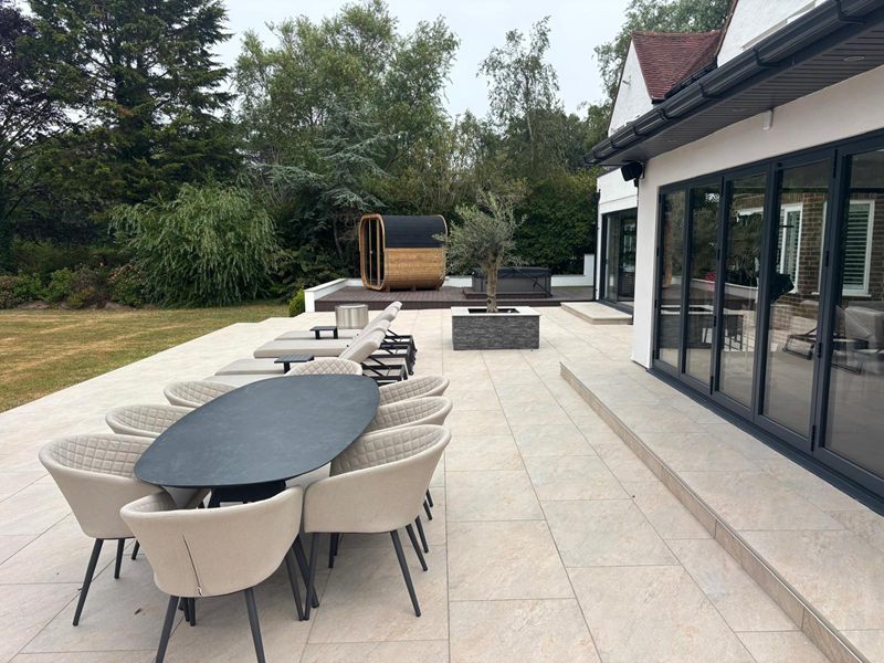 Large porcelain paved terrace with outdoor dining, sun loungers and a planted olive tree beside bi-fold doors