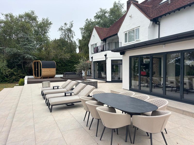 Wide view of a paved terrace with dining furniture and sun loungers, looking back towards the house