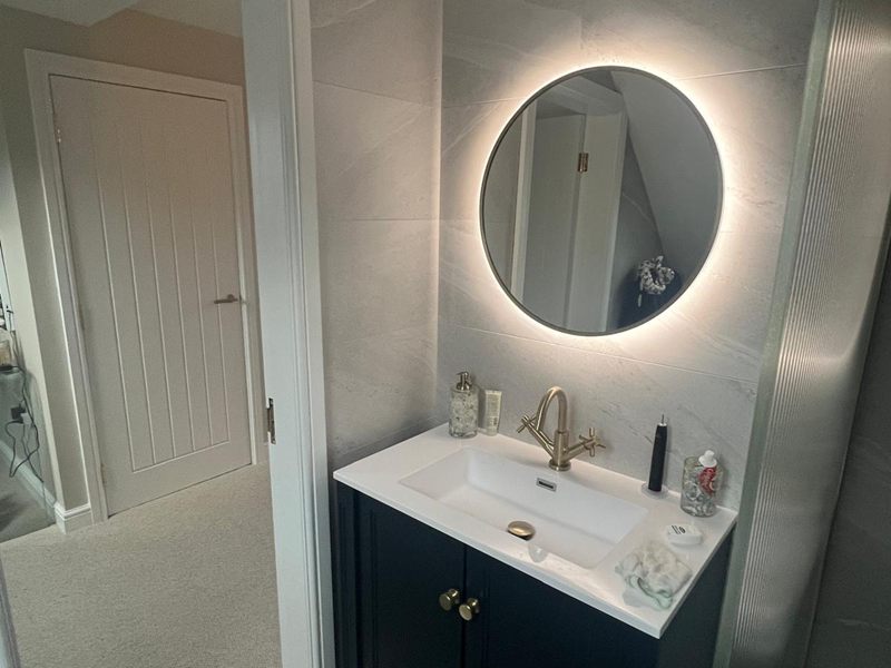 Loft en-suite with a navy vanity unit, brass tap and a backlit round mirror against marble effect tiling