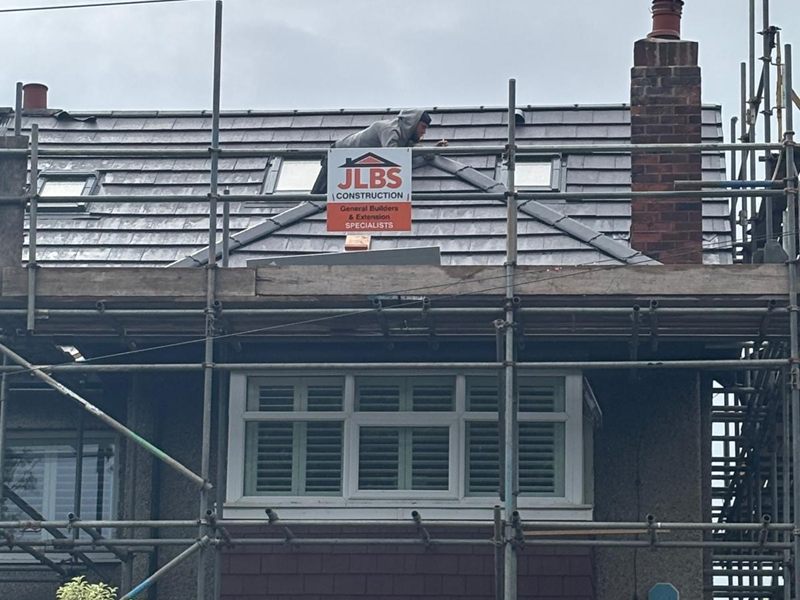 A roofer working on the new slate roof of a loft conversion beside a JLBS Construction site board